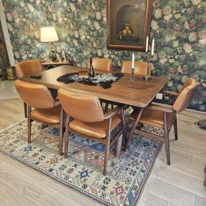 May include: A rectangular wooden dining table with rounded corners, paired with six tan leather chairs featuring dark wood frames. The table surface holds a black and white faux fur runner, a dark glass decanter, and two silver candle holders.