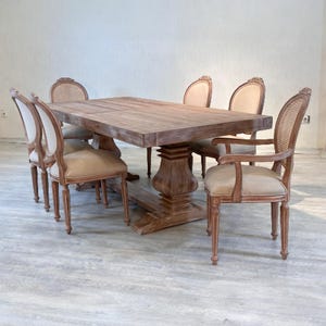 May include: A rectangular wooden dining table with a central pedestal base and six matching chairs. The chairs feature light beige upholstered seats and backrests with woven cane detailing on the back frames, supported by wooden legs.