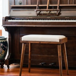 Puede incluir: Un taburete de piano de madera con un cojín rectangular de color crema. El taburete tiene cuatro patas cónicas y un marco de madera oscura. Un piano antiguo se encuentra en el fondo, con el texto "Farrell & Son London".