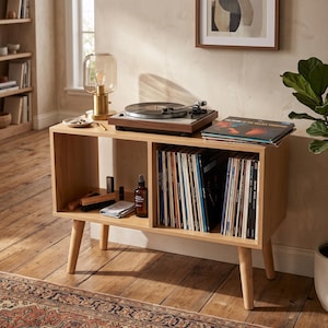 May include: A light wood record player console with four tapered legs, featuring two open storage compartments. The top surface holds a turntable, a glass lamp, and a stack of vinyl records. The lower shelves contain rows of vinyl records and accessories.