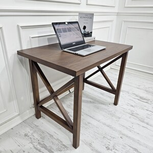 Puede incluir: Un escritorio de madera marrón oscuro con un diseño simple y moderno. El escritorio tiene una tapa rectangular y una base en forma de cruz. Un ordenador portátil está sobre el escritorio.