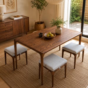 Puede incluir: Una mesa de comedor rectangular de madera oscura con cuatro patas y tres taburetes a juego con asientos tapizados en gris claro. La mesa sostiene un cuenco de madera con tres peras verdes y una pequeña taza gris oscuro sobre una alfombra.
