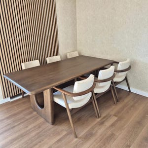 Puede incluir: Una mesa de comedor de madera oscura con una base curva única y seis sillas tapizadas blancas. La mesa está ubicada en una habitación con paneles de madera en una pared.