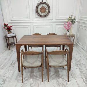 Könnte beinhalten: Ein Esstisch aus Holz mit vier Stühlen. Der Tisch ist braun und rechteckig. Die Stühle sind ebenfalls braun und haben ein schlichtes Design. Tisch und Stühle stehen vor einer weißen Wand.