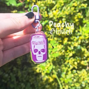 Könnte beinhalten: Ein klarer Acryl-Schlüsselanhänger mit einem lila Getränkedosen-Design mit einem Totenkopf-Motiv. Auf der Dose steht "Tero Tonic" und ist an einem silberfarbenen Verschluss befestigt. Der Hintergrund ist verschwommenes grünes Laub.