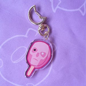 Könnte beinhalten: Ein rosa schlüsselförmiger Anhänger in Eisform mit einem Totenkopf-Design. Der Schlüsselanhänger hat einen goldfarbenen Verschluss und einen transparenten Acrylkörper. Der Schädel hat ein schwarzes Auge und einen traurigen Ausdruck. Der Eisstiel ist hellbraun.