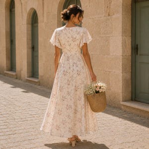 May include: A floor-length cream-colored dress with a delicate floral pattern, featuring short flutter sleeves, a fitted bodice, and a flowing skirt. The dress is paired with a woven straw basket containing a bouquet of light pink flowers.