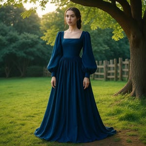 Op de afbeelding: Een vrouw draagt een lange, donkerblauwe jurk met een vierkante halslijn en lange, pofmouwen. De jurk heeft een volle rok en bevindt zich in een groene buitenomgeving met een boom en een houten hek op de achtergrond.