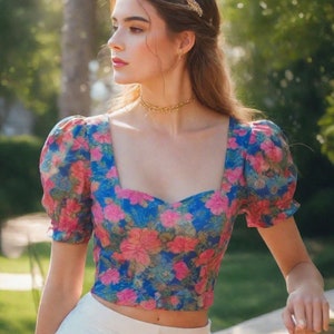 Puede incluir: Una mujer con un top corto de flores azules y rosas con mangas abullonadas y pantalones blancos. Está de pie al aire libre con un fondo borroso de árboles verdes.