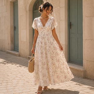May include: A floor-length cream-colored dress featuring a delicate pink and green floral pattern, a deep V-neckline, and flutter sleeves. The dress has a fitted bodice that transitions into a flowing, ankle-length skirt, paired with a woven straw bag.