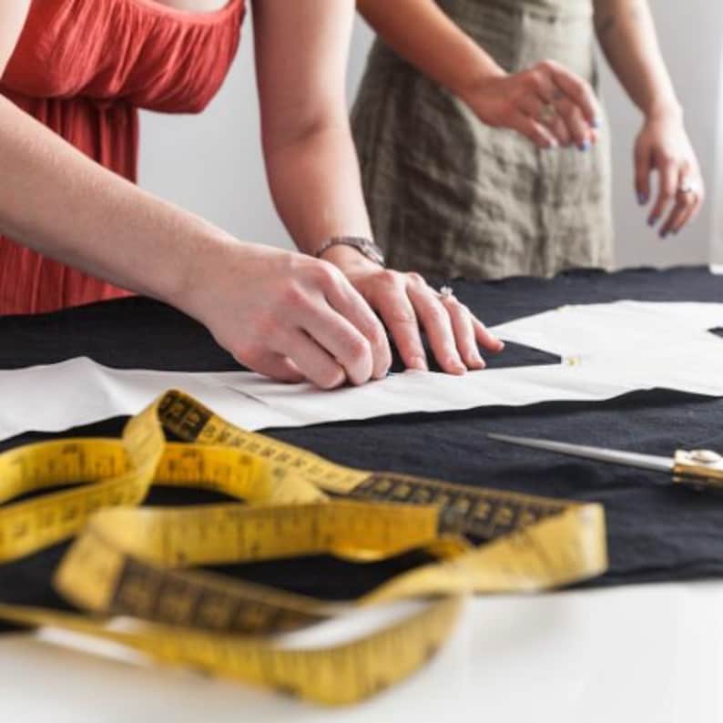 Op de afbeelding: Twee mensen werken aan een naaiproject. E&eacute;n persoon gebruikt een potlood om een patroon te tekenen op witte stof die op een zwart oppervlak ligt. Een gele meetlint en een schaar staan op de voorgrond.