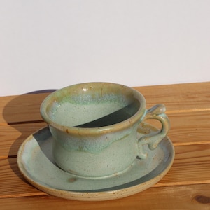 Könnte beinhalten: Ein handgefertigtes Keramik-Tassen- und Untertassen-Set. Die Tasse und die Untertasse sind in einem weichen, gedämpften Grün gehalten und haben eine gesprenkelte, strukturierte Oberfläche. Der Rand der Tasse hat eine hellbraune Glasur.