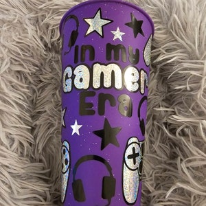 May include: Purple reusable cup with a glittery finish. The cup has a black design with the text "In my gamer Era" and images of video game controllers and headphones.