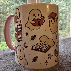 May include: A ceramic mug with a pale pink handle and rim. The mug displays a pattern of cartoon ghosts reading books, with stars, leaves, and a crescent moon. The ghosts are in different poses, some sitting on stacks of books.