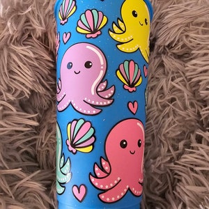 May include: A blue reusable cup with a glitter finish featuring a pattern of pink, yellow, and green octopuses with white dots and seashells. The cup has a white rim and a white base.