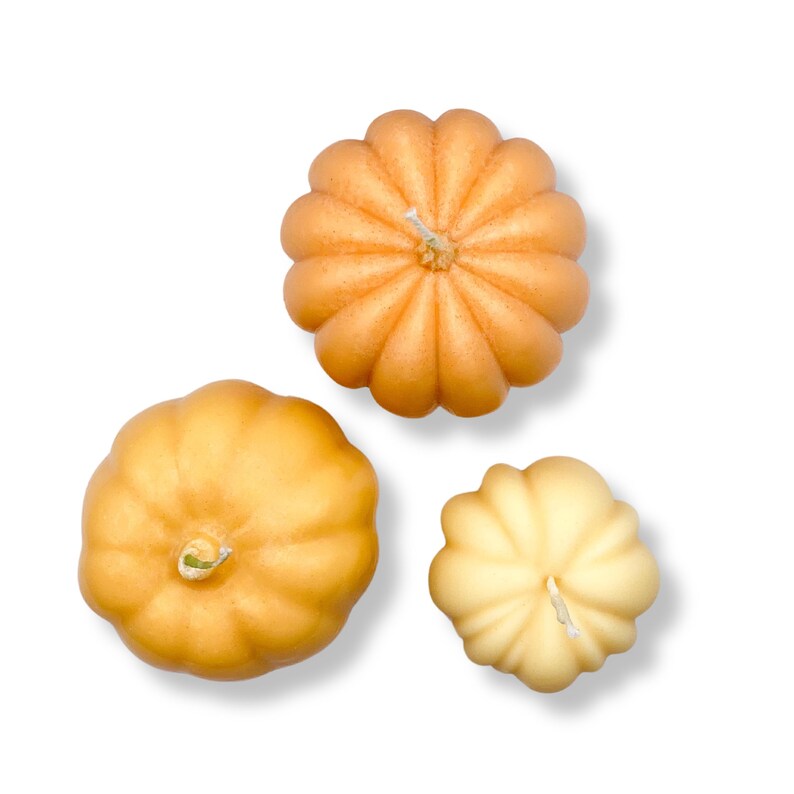 Puede incluir: Tres velas decorativas con forma de calabaza en tonos naranja y amarillo. Las velas tienen una textura acanalada y est&aacute;n dispuestas sobre un fondo blanco.