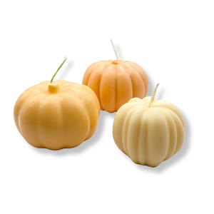 Puede incluir: Tres velas con forma de calabaza, una naranja, una amarilla y una crema. Las velas tienen todas la forma de una calabaza con un tallo en la parte superior.
