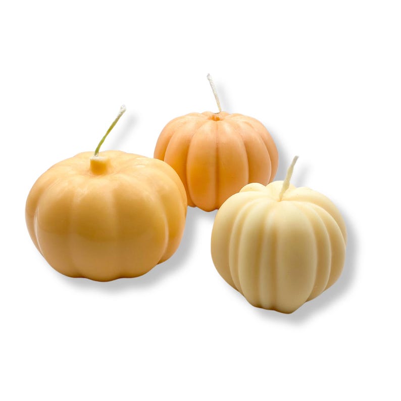 Puede incluir: Tres velas con forma de calabaza, una naranja, una amarilla y una crema. Las velas tienen todas la forma de una calabaza con un tallo en la parte superior.