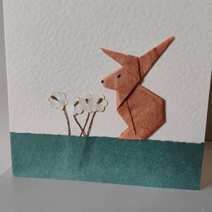 May include: A handmade greeting card with a brown origami rabbit and three white paper flowers. The rabbit is on a white background above a teal green strip. The card is a unique, artistic design, perfect for Easter.
