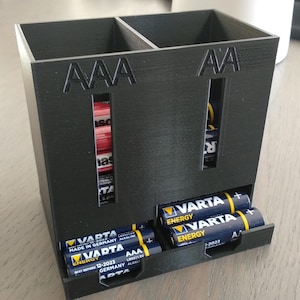 May include: Black battery storage unit, designed to hold AAA and AA batteries. The unit has visible compartments and the text "AAA" and "AA". Several batteries are visible, including Varta brand batteries.