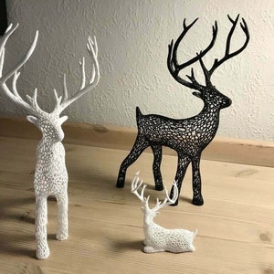 Op de afbeelding: Drie decoratieve rendierfiguren, één zwart en twee wit, met ingewikkelde, kant-achtige ontwerpen. De rendieren staan op een houten oppervlak.