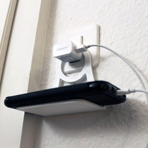 May include: A white 3D printed phone holder with a shelf for a phone and a built-in charging port. A white phone charger is plugged into the wall outlet and a white charging cable is connected to the phone.