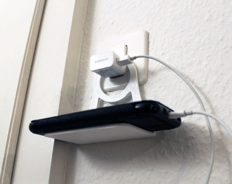 K&ouml;nnte beinhalten: Ein wei&szlig;er 3D-gedruckter Telefonhalter mit einem Regal f&uuml;r ein Telefon und einem integrierten Ladeanschluss. Ein wei&szlig;es Telefonladeger&auml;t ist in die Steckdose gesteckt und ein wei&szlig;es Ladekabel ist mit dem Telefon verbunden.