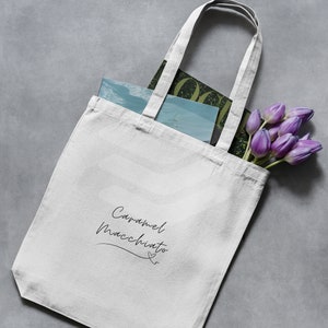 Puede incluir: Bolsa de tela blanca con el texto "Caramel Macchiato" escrito en cursiva negra. La bolsa está llena de una revista y un ramo de tulipanes morados.