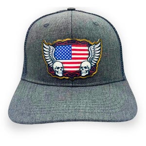 May include: A gray trucker hat with a mesh back and a curved brim. The front features a patch with the American flag, white wings, and two skulls. The patch has a gold-colored border.