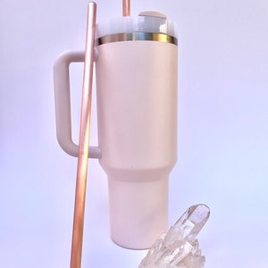 May include: A pink stainless steel tumbler with a lid and a handle. The tumbler has two copper straws sticking out of the lid. A clear quartz crystal cluster is in front of the tumbler.
