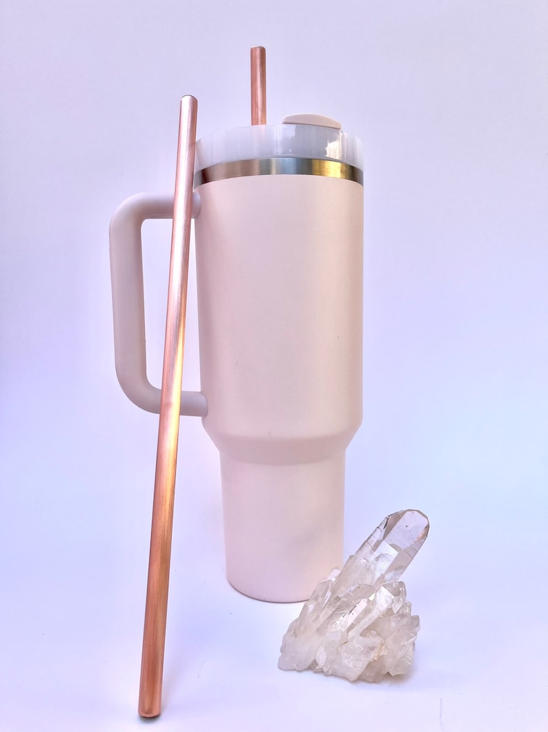 May include: A pink stainless steel tumbler with a lid and a handle. The tumbler has two copper straws sticking out of the lid. A clear quartz crystal cluster is in front of the tumbler.