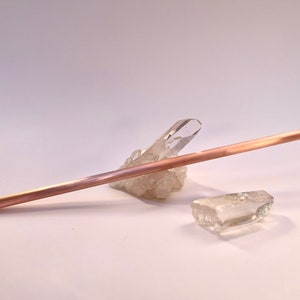 May include: A rose gold metal straw resting on a cluster of clear quartz crystals.