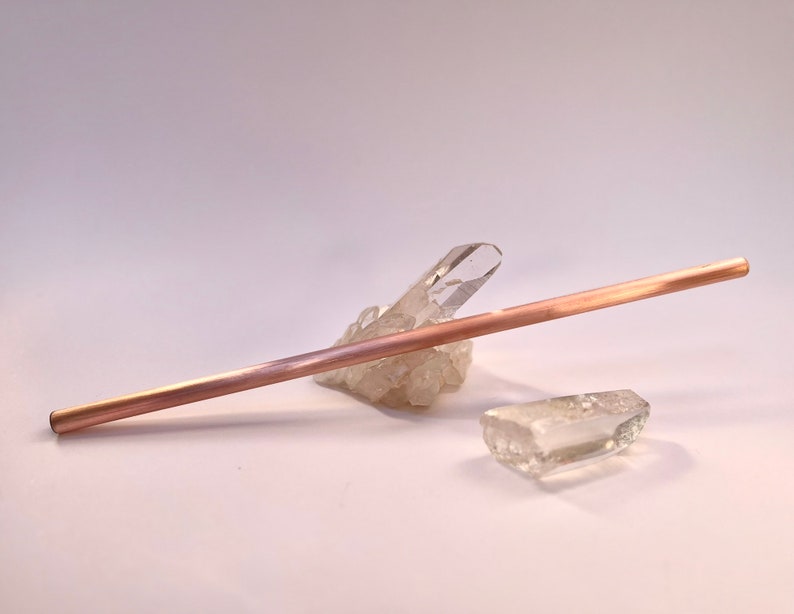 May include: A rose gold metal straw resting on a cluster of clear quartz crystals.