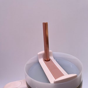 May include: A white travel mug with a stainless steel band and a pink lid. The lid has a built-in straw holder and a copper straw is inserted into the holder.