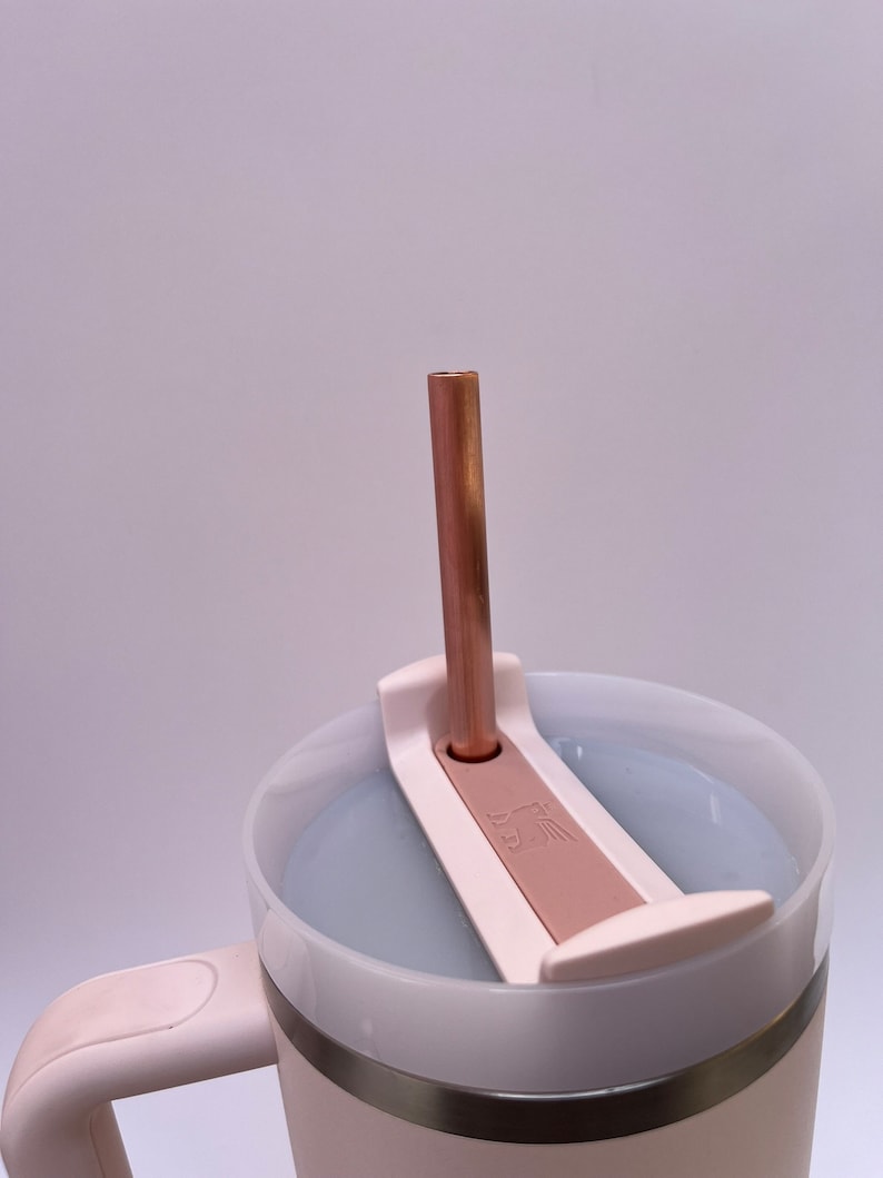 May include: A white travel mug with a stainless steel band and a pink lid. The lid has a built-in straw holder and a copper straw is inserted into the holder.