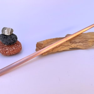May include: A rose gold metal drinking straw resting on a piece of driftwood.  The straw is long and thin.  There are three small rocks stacked on a white surface in the background.