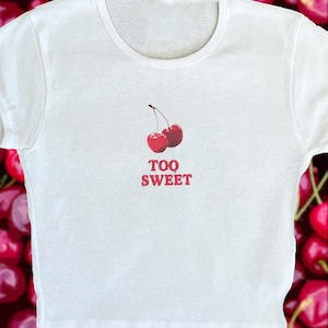 May include: White t-shirt with a red graphic of two cherries and the text "TOO SWEET" printed on the front.
