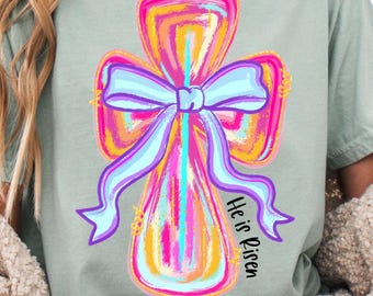 Op de afbeelding: Een lichtgroen T-shirt met een kleurrijk kruisontwerp en een strik. Het kruis is geschilderd in tinten roze, oranje, geel en blauw. De tekst "He is Risen" staat aan de zijkant van het kruis.