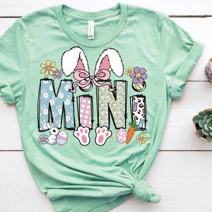 May include: A light green t-shirt with a graphic design of the word "MINI" in a patchwork style. The design features bunnies, eggs, carrots, and flowers.