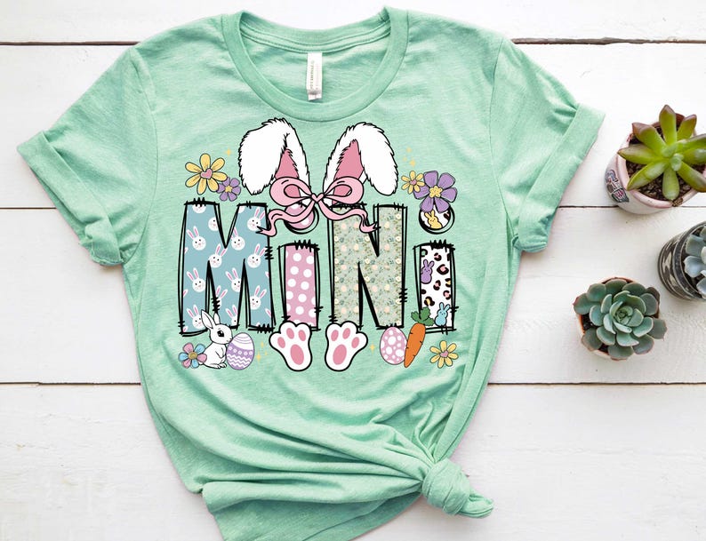 May include: A light green t-shirt with a graphic design of the word "MINI" in a patchwork style. The design features bunnies, eggs, carrots, and flowers.