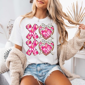 以下が含まれることがあります： 前面にピンクと黒のハートのデザインと「XO XO」の文字がプリントされた白いTシャツです。