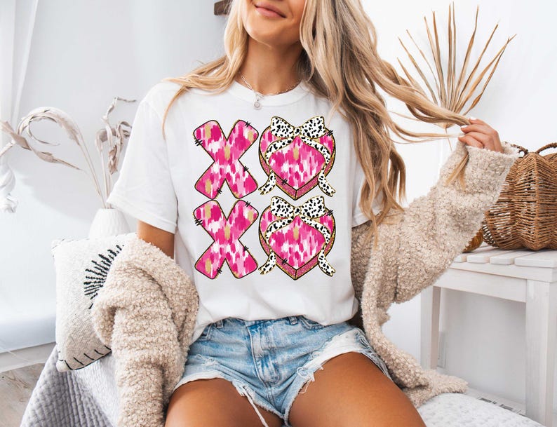 以下が含まれることがあります： 前面にピンクと黒のハートのデザインと「XO XO」の文字がプリントされた白いTシャツです。