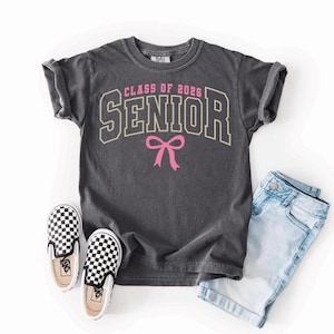 May include: A charcoal gray t-shirt with the text "CLASS OF 2026 SENIOR" in a collegiate style font, with a pink bow graphic. The shirt is paired with light blue denim shorts and black and white checkered slip-on sneakers.