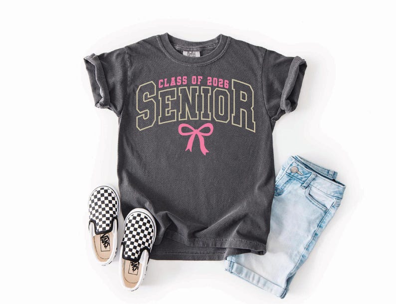 May include: A charcoal gray t-shirt with the text "CLASS OF 2026 SENIOR" in a collegiate style font, with a pink bow graphic. The shirt is paired with light blue denim shorts and black and white checkered slip-on sneakers.