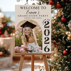 Pode incluir: Um cavalete de madeira com um letreiro de boas-vindas para uma festa de formatura. O letreiro diz "Bem-vindo &agrave; festa de formatura de Emma, Lincoln High School, Turma de 2026". Uma foto de uma graduada sorrindo &eacute; apresentada.