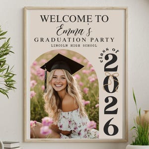 Pode incluir: Uma placa emoldurada para uma festa de formatura com o texto "Welcome to Emma's Graduation Party" e "Class of 2026". A placa apresenta uma foto de uma graduada sorrindo usando um chap&eacute;u e uma beca, cercada por flores cor-de-rosa.