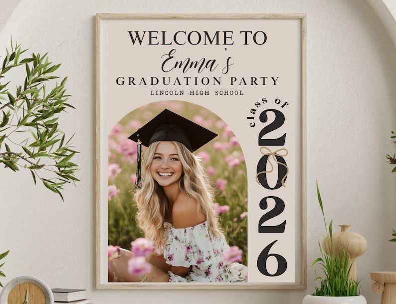 Pode incluir: Uma placa emoldurada para uma festa de formatura com o texto "Welcome to Emma's Graduation Party" e "Class of 2026". A placa apresenta uma foto de uma graduada sorrindo usando um chap&eacute;u e uma beca, cercada por flores cor-de-rosa.
