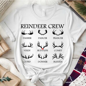 May include: White t-shirt with the words "REINDEER CREW" and illustrations of reindeer antlers. Each antler design is labelled with a reindeer name: Dasher, Dancer, Prancer, Vixen, Rudolph, Comet, Cupid, Donner, and Blitzen. A festive holiday theme.