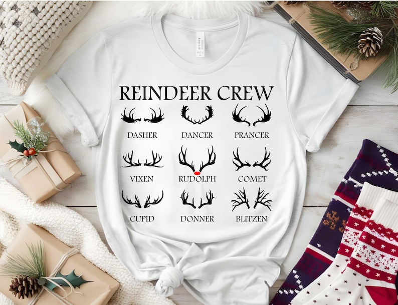 May include: White t-shirt with the words "REINDEER CREW" and illustrations of reindeer antlers. Each antler design is labelled with a reindeer name: Dasher, Dancer, Prancer, Vixen, Rudolph, Comet, Cupid, Donner, and Blitzen. A festive holiday theme.