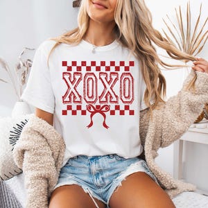 以下が含まれることがあります： 赤いチェック柄の白いTシャツに、赤い「XOXO」の文字と、その下に赤いリボンが描かれています。